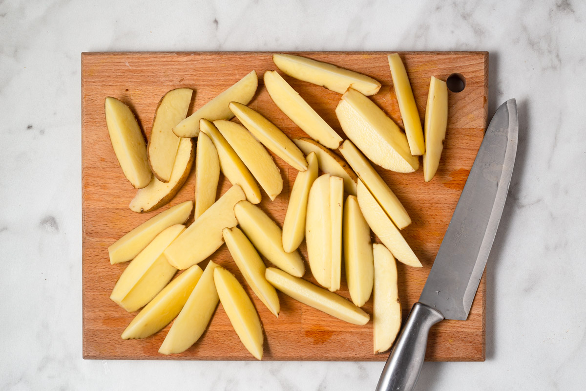 potatoes cut into wedges on a chopping board with a knife.