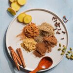 Chai Spice mix ingredients on a plate with ginger and cinnamon sticks on the side.