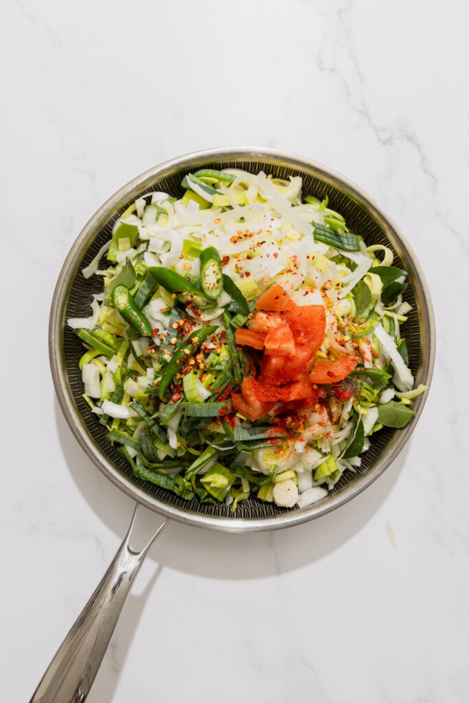 Chopped leeks, green chilies, onions, and tomatoes with spices in a pan on a white background.