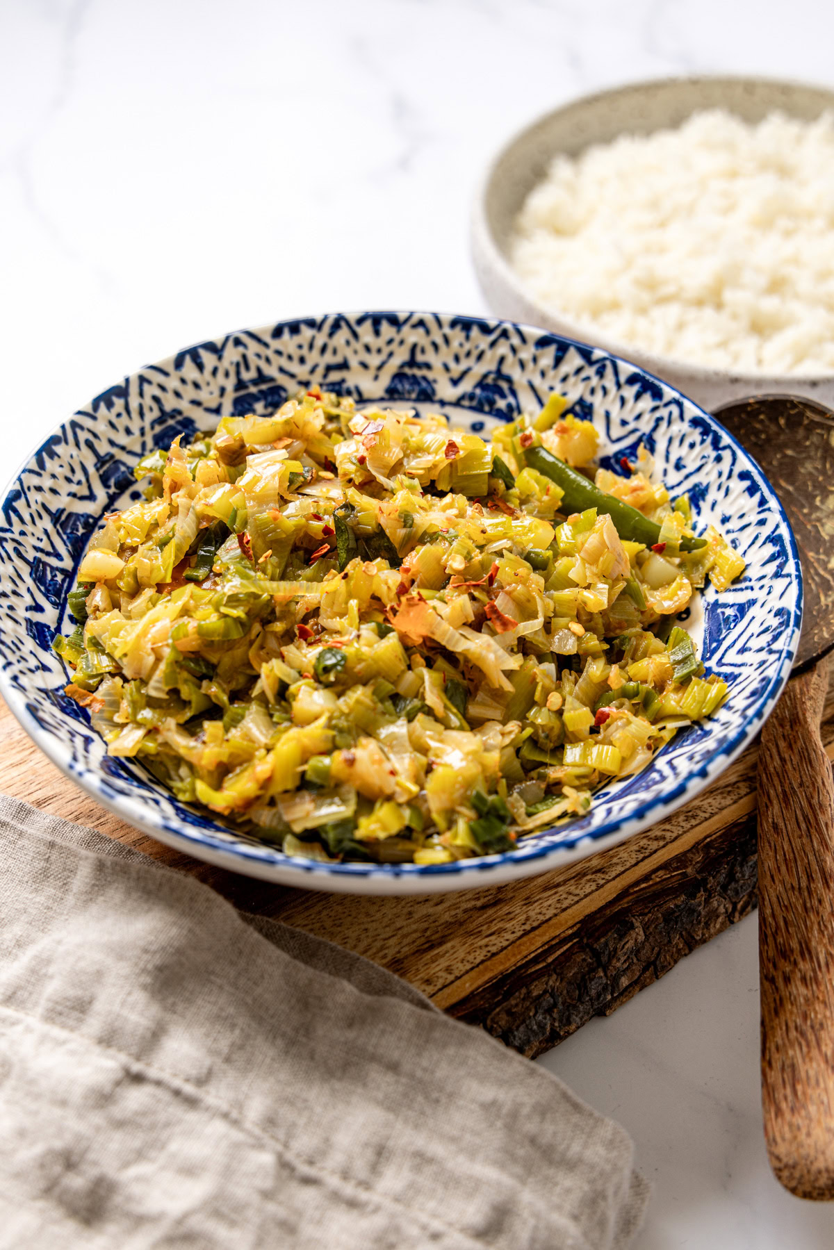 Bowl of Sri Lankan leeks curry on a wooden board beside rice and a wooden spoon.
