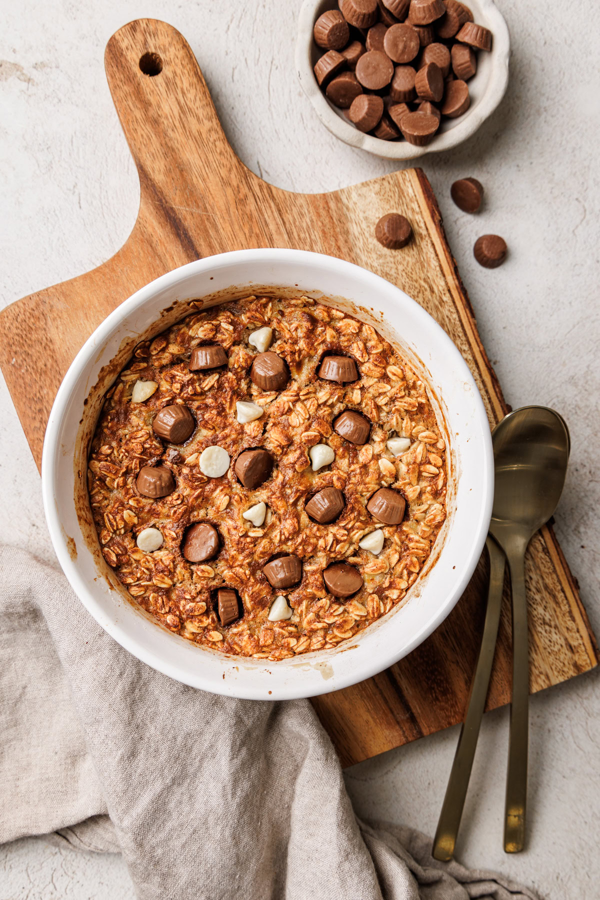 Freshly baked oats with chocolate pieces and white chocolate chips in a white dish, set on a wooden cutting board against a light stone-textured background.