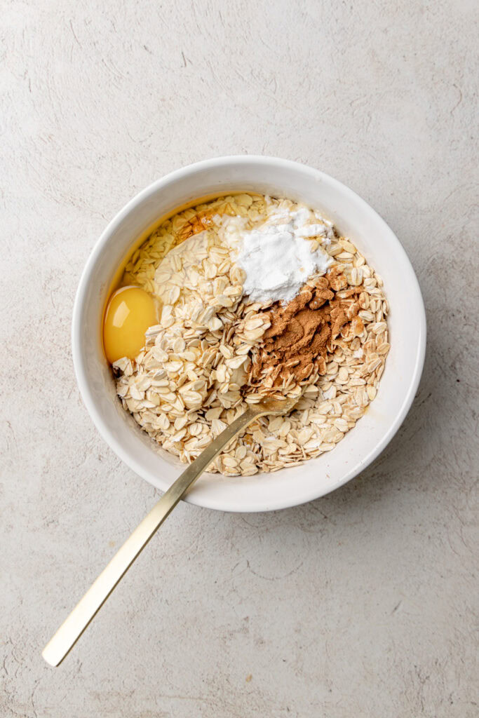 Rolled oats, egg, yogurt, and cinnamon in a white bowl on a light stone-textured surface before mixing.