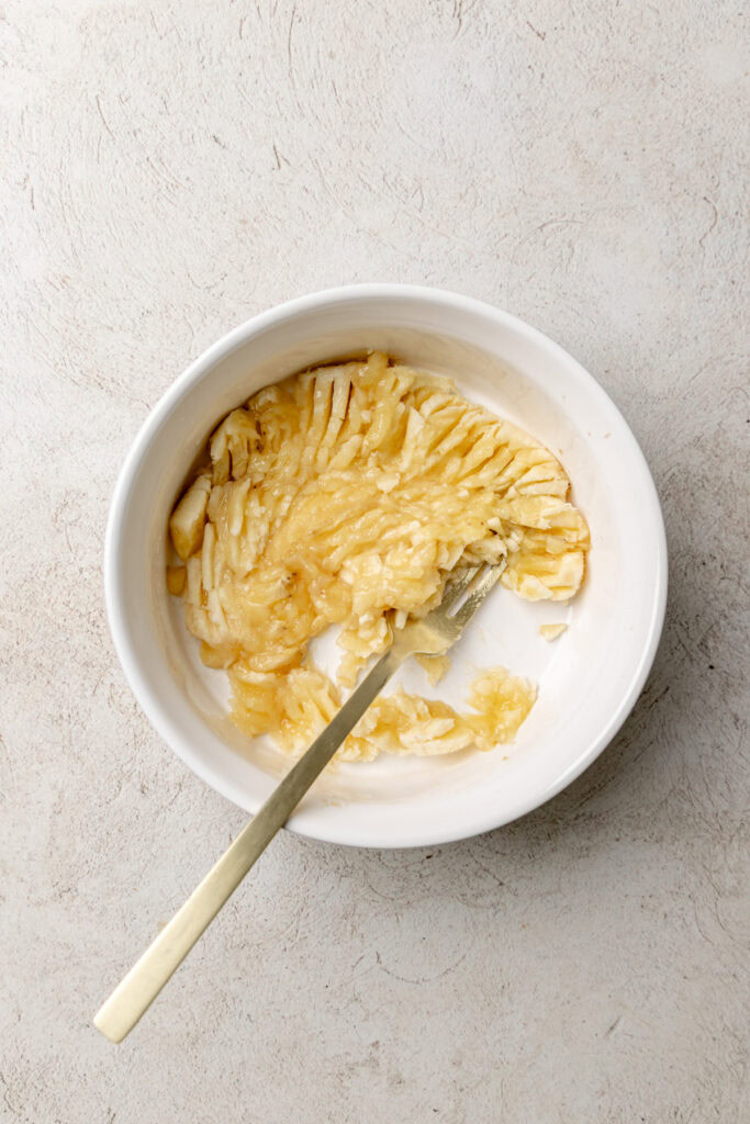 Mashed banana in a white bowl with a fork, set on a light stone-textured surface.